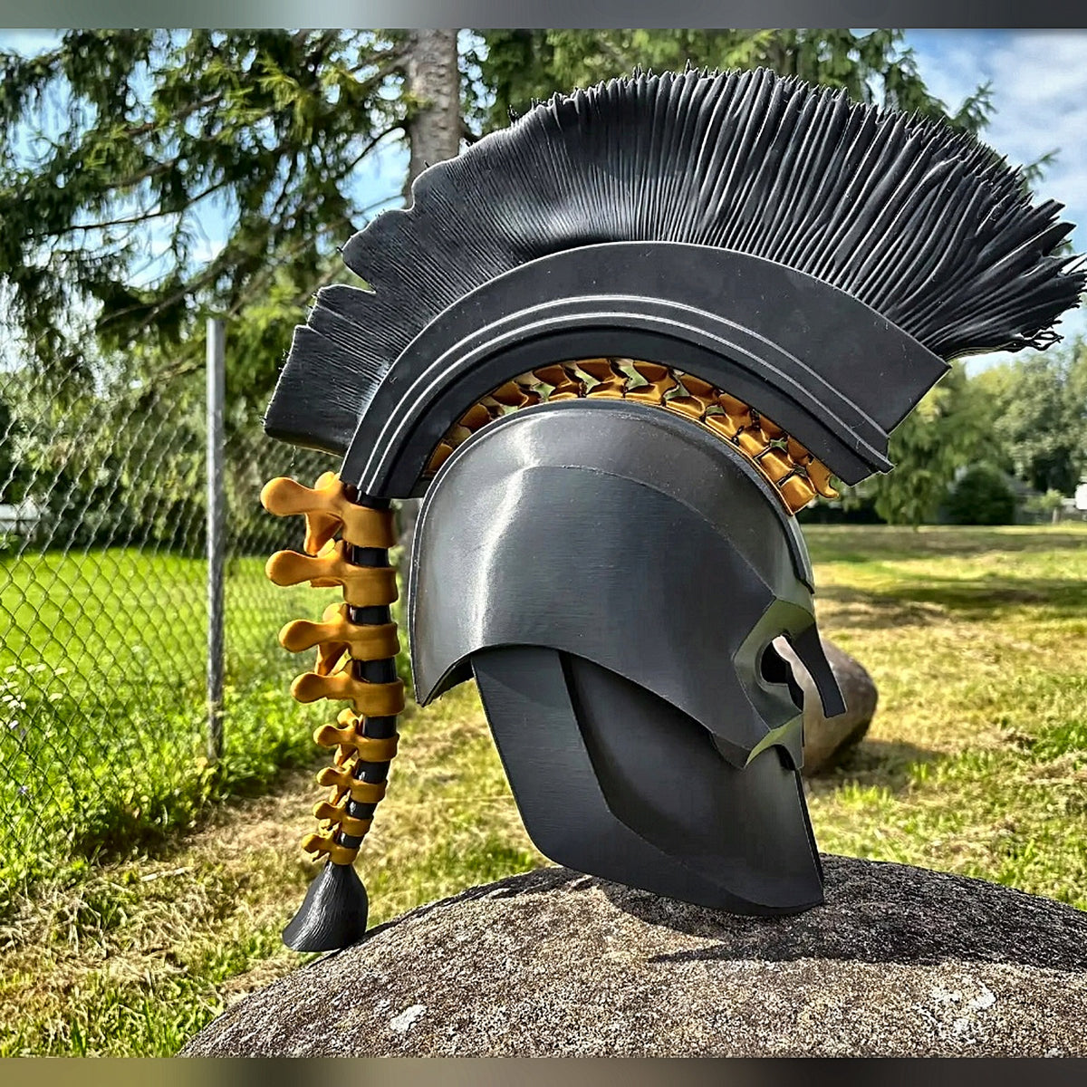 Black Greek warrior helmet with tall black crest and gold spine, outdoors on a rock